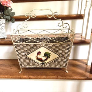 May include: A decorative magazine rack with a woven basket design and a rooster emblem. The rack has a cream-colored metal frame with decorative scrollwork and four small feet. The rooster emblem is in a diamond shape.