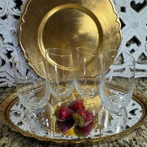 May include: Four clear glass tumblers with a textured surface sit on a gold mirrored tray with a decorative border. The tray is surrounded by a white carved wooden surface and a gold charger plate.