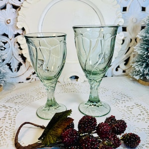 May include: Two green glass wine glasses with a textured design. The glasses are on a white lace tablecloth with a sprig of red berries.