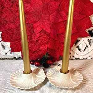May include: Two gold taper candles in off-white, shell-shaped candle holders. The candles stand in front of a red poinsettia decoration. The candle holders have a decorative leaf pattern.