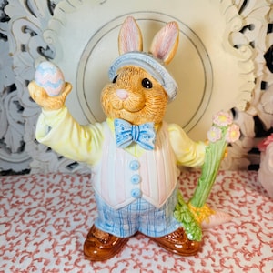 May include: A ceramic Easter bunny figurine, wearing a blue plaid suit, a striped vest, and a light blue hat. The bunny holds a pastel-colored egg in one hand and a flower-topped carrot in the other. The figurine has brown shoes and a blue bow tie.
