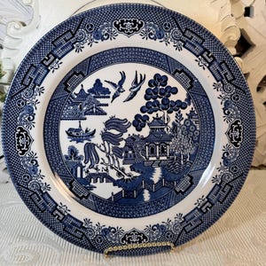 May include: A blue and white porcelain dinner plate with a traditional willow pattern. The plate features a detailed design of a bridge, a pagoda, a willow tree, and birds. The plate is on a white stand.