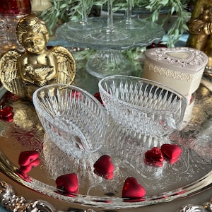 May include: A silver serving tray holds two clear, cut-glass dishes, red heart-shaped candies, and a gold cherub statue. A heart-shaped trinket box and a glass cake stand with champagne flutes are in the background. The scene has a romantic, vintage aesthetic.