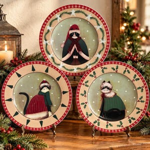 May include: Three round ceramic plates featuring illustrations of cats dressed in winter clothing. Each plate has a cream-colored rim with red borders and holiday-themed patterns like stars, pine trees, and holly leaves.