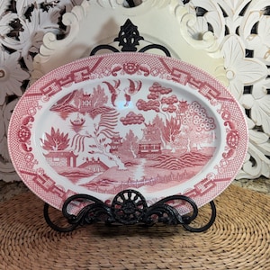 May include: An oval ceramic platter featuring a red and white transferware pattern depicting a landscape with pagodas, trees, a bridge, and two birds. The platter is displayed on a black metal stand, showcasing a detailed scene with intricate borders.