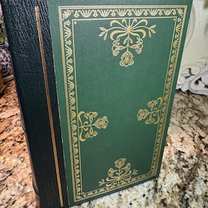 May include: A hardcover book with a green cover and gold floral designs. The spine is black with a gold accent. The book is standing upright, showcasing its decorative cover.