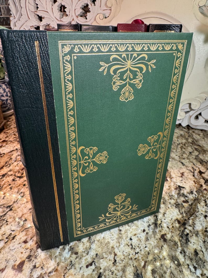 May include: A hardcover book with a green cover and gold floral designs. The spine is black with a gold accent. The book is standing upright, showcasing its decorative cover.