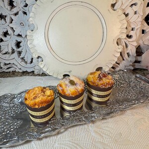 May include: Three small, round, sweet bread muffins with brown paper wrappers. The muffins are on a silver tray with an ornate design. The tray is on a white surface.