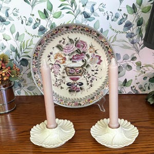 May include: Two cream-colored ceramic candle holders with a scalloped, shell-like base design, each holding a tall, smooth, light pink taper candle. The candle holders are positioned in front of a decorative ceramic plate featuring a floral pattern.