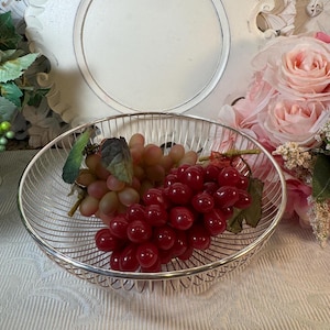 May include: A silver wire fruit bowl with two bunches of grapes, one red and one green, inside. The bowl is on a white surface.