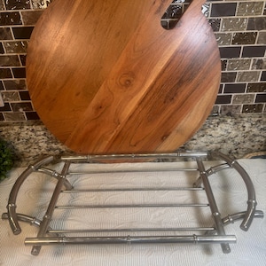 May include: A round wooden cutting board with a handle is displayed behind a silver-colored metal serving rack. The cutting board is made of light brown wood with a natural grain pattern. The serving rack has a bamboo-like design.