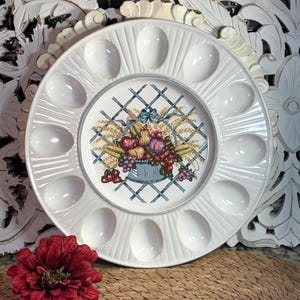 May include: A round white ceramic egg serving platter featuring twelve oval indentations arranged in a circle around a central decorative motif. The center displays a colorful illustration of a basket filled with various fruits and wheat stalks.