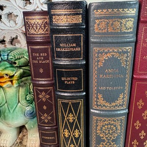 May include: A collection of antique books with decorative gold accents. The spines are various colors, including burgundy, black, and gray. Titles include "The Red and the Black," "William Shakespeare Selected Plays," and "Anna Karenina."