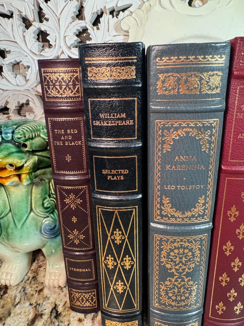 May include: A collection of antique books with decorative gold accents. The spines are various colors, including burgundy, black, and gray. Titles include "The Red and the Black," "William Shakespeare Selected Plays," and "Anna Karenina."