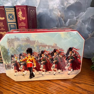 May include: Vintage tin box with a red trim and a colorful illustration of a Scottish military band marching in front of a castle. The band members wear red and black kilts and play bagpipes. The box is rectangular with an octagonal shape.