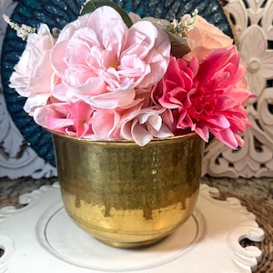 May include: A gold-colored, hammered metal vase filled with pink and red flowers. The vase sits on a white, ornate, scalloped-edge plate. The background includes a teal, textured wall hanging and a woven placemat.