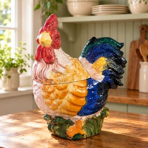 May include: A ceramic rooster-shaped jar with a removable lid. The rooster features a bright red comb and wattle, a yellow beak, and a body with white, orange, and blue feathers. The base is sculpted to resemble green grass with orange feet.