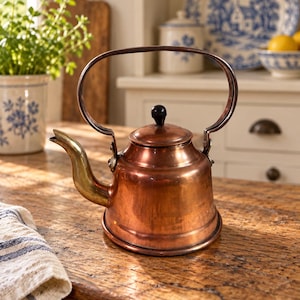 May include: A polished copper tea kettle with a curved brass spout and a tall, arched handle. The kettle features a rounded body and a small, dark knob on the lid. The metal surface reflects light, showing warm orange and brown metallic tones.