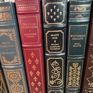 May include: A collection of antique books with gold lettering and decorative accents. Titles include "Anna Karenina," "The Three Musketeers," "Vanity Fair," and "Wuthering Heights." The books have leather-bound covers in various colors, including gray, burgundy, and black.