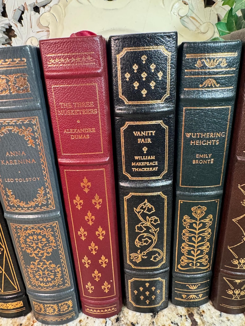 May include: A collection of antique books with gold lettering and decorative accents. Titles include "Anna Karenina," "The Three Musketeers," "Vanity Fair," and "Wuthering Heights." The books have leather-bound covers in various colors, including gray, burgundy, and black.