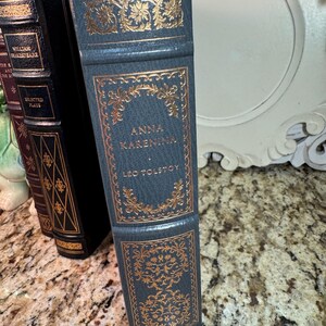 May include: A stack of antique books with gold lettering and accents. The spine of the front book is teal with the title "Anna Karenina" by Leo Tolstoy. Other books are visible with black and burgundy covers.