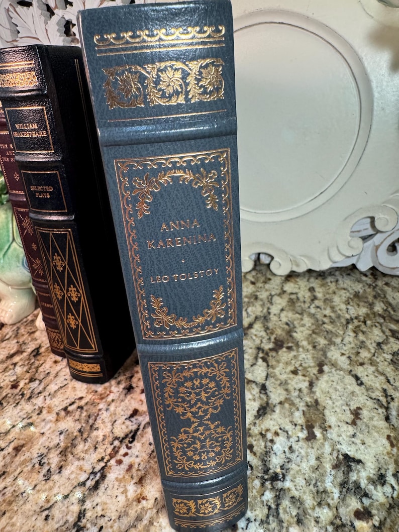 May include: A stack of antique books with gold lettering and accents. The spine of the front book is teal with the title "Anna Karenina" by Leo Tolstoy. Other books are visible with black and burgundy covers.