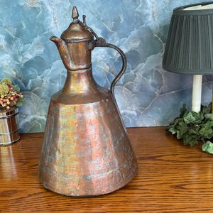 May include: A tall, conical copper pitcher featuring a weathered, multi-toned metallic finish with visible horizontal striations. It includes a narrow spout, a curved handle, and a pointed lid attached by a small metal hinge at the top.