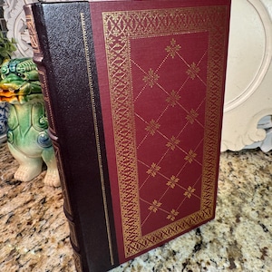 May include: A hardcover book with a burgundy cover and a dark brown spine. The burgundy cover features a gold geometric pattern. The spine is dark brown with gold lettering. The book is displayed at an angle.