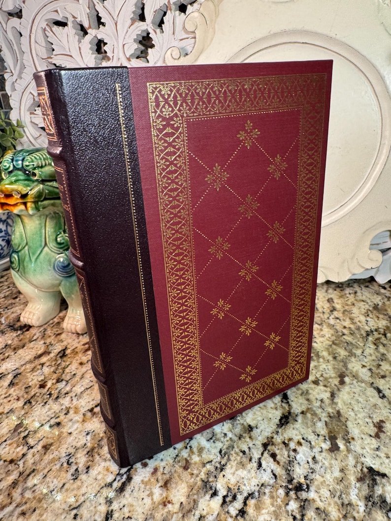 May include: A hardcover book with a burgundy cover and a dark brown spine. The burgundy cover features a gold geometric pattern. The spine is dark brown with gold lettering. The book is displayed at an angle.