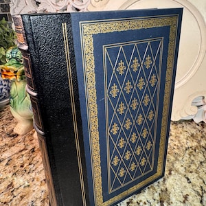 May include: A hardcover book with a black spine and a navy blue cover. The cover features a gold fleur-de-lis pattern within a gold border. The book's edges are also gold, and the spine has gold accents.