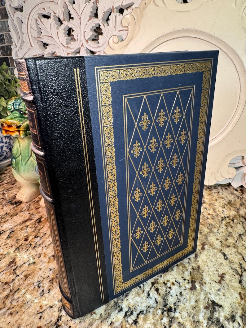 May include: A hardcover book with a black spine and a navy blue cover. The cover features a gold fleur-de-lis pattern within a gold border. The book's edges are also gold, and the spine has gold accents.