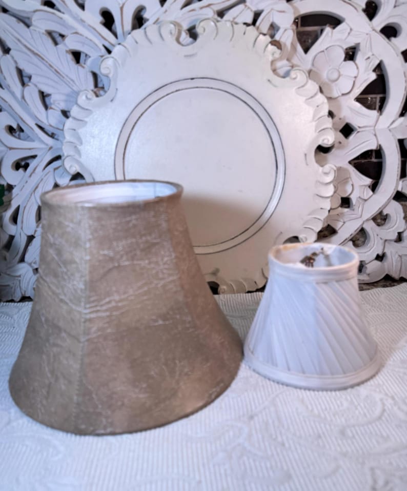 May include: Two lampshades, one is a larger beige shade with a textured surface and the other is a smaller white shade with a pleated design.