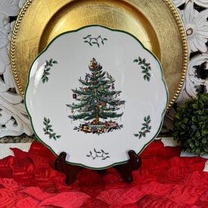 May include: A white ceramic Christmas plate with a green scalloped edge. The plate features a detailed illustration of a decorated Christmas tree with presents underneath. Sprigs of holly are placed around the edge.
