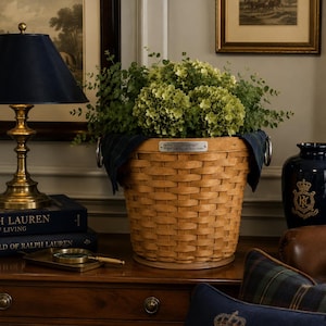 May include: A woven light brown wooden basket with a silver metal plaque, filled with green hydrangea flowers and foliage. The basket features two silver metal side handles and is positioned between a brass lamp and a dark blue ceramic vase.