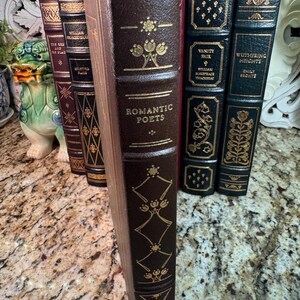 May include: A collection of antique books with dark brown leather covers and gold lettering. The spine of the book in the foreground reads "Romantic Poets." Other titles include "Vanity Fair" and "Wuthering Heights."