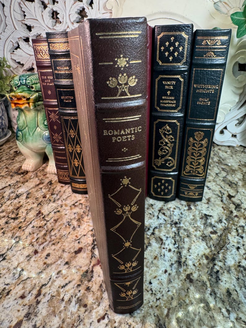 May include: A collection of antique books with dark brown leather covers and gold lettering. The spine of the book in the foreground reads "Romantic Poets." Other titles include "Vanity Fair" and "Wuthering Heights."