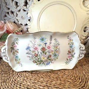 May include: Rectangular white ceramic serving tray with a floral design in the center and on the sides. The flowers are in shades of pink, blue, yellow, and purple. The tray has a gold trim and decorative handles.