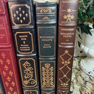 May include: A collection of classic books with decorative gold lettering and accents. Titles include "Vanity Fair," "Wuthering Heights," and "Romantic Poets." The books have leather-bound covers in various colors, including black, green, and brown.