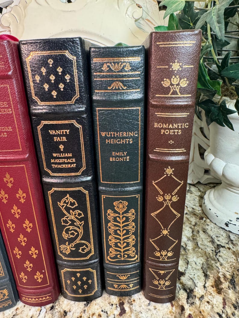 May include: A collection of classic books with decorative gold lettering and accents. Titles include "Vanity Fair," "Wuthering Heights," and "Romantic Poets." The books have leather-bound covers in various colors, including black, green, and brown.