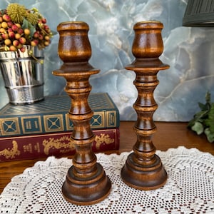 May include: Two vintage wooden candle holders with a rich, dark brown finish. The candle holders have a classic, turned design with a wide base and a flared top. They are displayed on a white lace doily, suggesting a rustic or antique aesthetic.