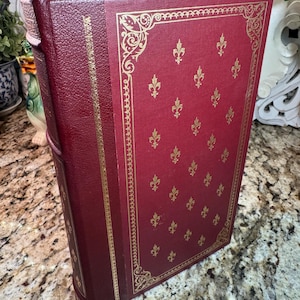 May include: A burgundy-colored book with gold fleur-de-lis pattern and gold trim. The book has a matching burgundy ribbon bookmark. The cover is embossed with a decorative gold border.