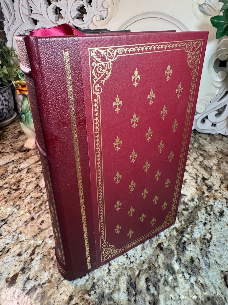 May include: A burgundy-colored book with gold fleur-de-lis pattern and gold trim. The book has a matching burgundy ribbon bookmark. The cover is embossed with a decorative gold border.
