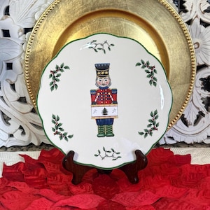 May include: A white ceramic plate with a green scalloped edge and a painted nutcracker soldier design. The plate is decorated with sprigs of holly and rests on a wooden stand, with a gold charger plate in the background. The plate is approximately 10 inches in diameter.