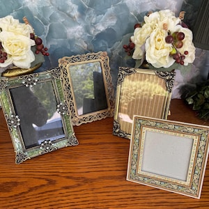 May include: A collection of four decorative picture frames. One frame is rectangular with green enamel and clear rhinestones. Another is an ornate gold-tone rectangular frame. A third is a gold-tone rectangular frame, and the fourth is square.