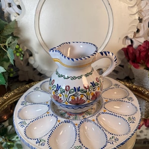 May include: A white ceramic egg plate with blue and green trim and floral designs. The plate has 10 individual egg-shaped indentations. A matching white ceramic pitcher with blue and green trim and floral designs sits in the center of the plate.