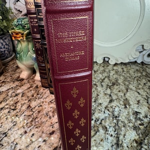 May include: A burgundy-colored book titled "The Three Musketeers" by Alexandre Dumas. The book has gold lettering and decorative gold fleur-de-lis patterns. The spine has gold lettering and a textured design.