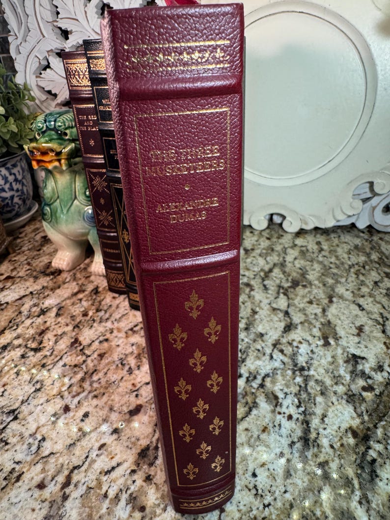 May include: A burgundy-colored book titled "The Three Musketeers" by Alexandre Dumas. The book has gold lettering and decorative gold fleur-de-lis patterns. The spine has gold lettering and a textured design.