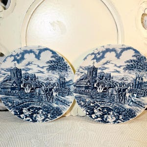 May include: Two blue and white decorative plates with a transferware design. Each plate features a horse-drawn carriage, a church, and a bridge. The plates have a classic, vintage aesthetic.