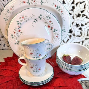 May include: A white porcelain dinnerware set with a green trim and a holly berry design. The set includes plates, bowls, cups, and saucers. Two cups are stacked on top of saucers. A stack of bowls contains two strawberries.