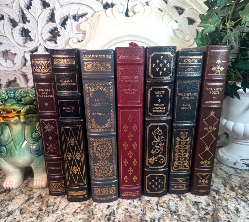 May include: A collection of antique books with decorative gold lettering and designs on the spines. The books are in various colors, including burgundy, black, and brown. Titles include "Anna Karenina" and "Wuthering Heights."